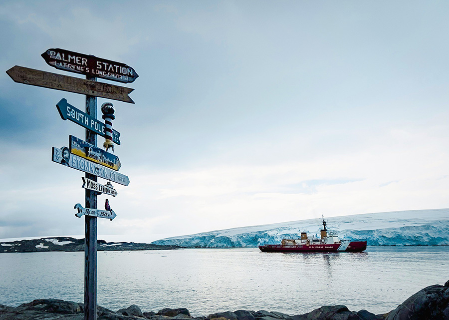 A ship sits in a harbor with a glacier behind it and a signpost in the foreground. 