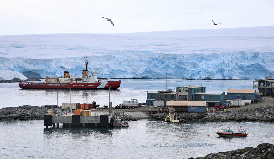 A ship sits in a harbor behind several buildings with a glacier in the background and birds circling. 