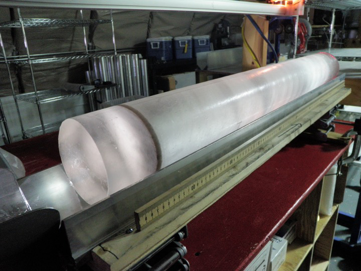 A man in a red jacket examines a long column of ice sitting horizontally on a table