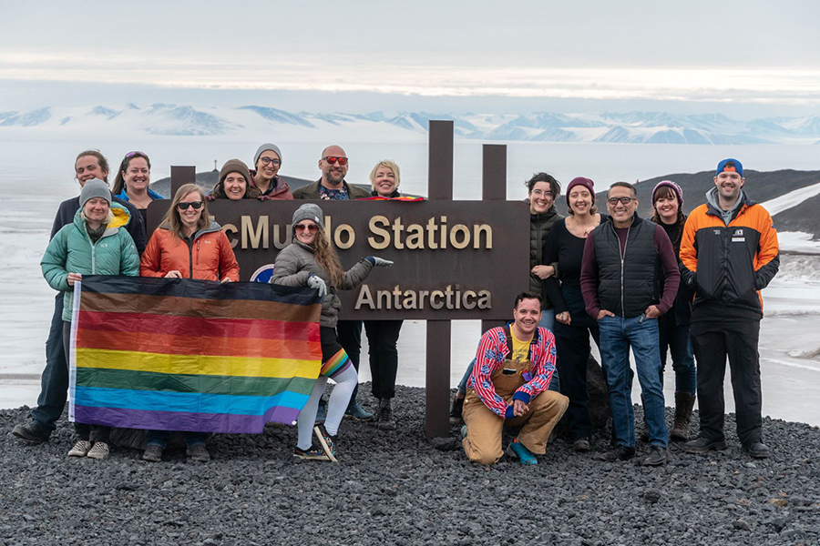 McMurdo Station staff and scientists pose with a pride flag in front of the McMurdo Station sign. 